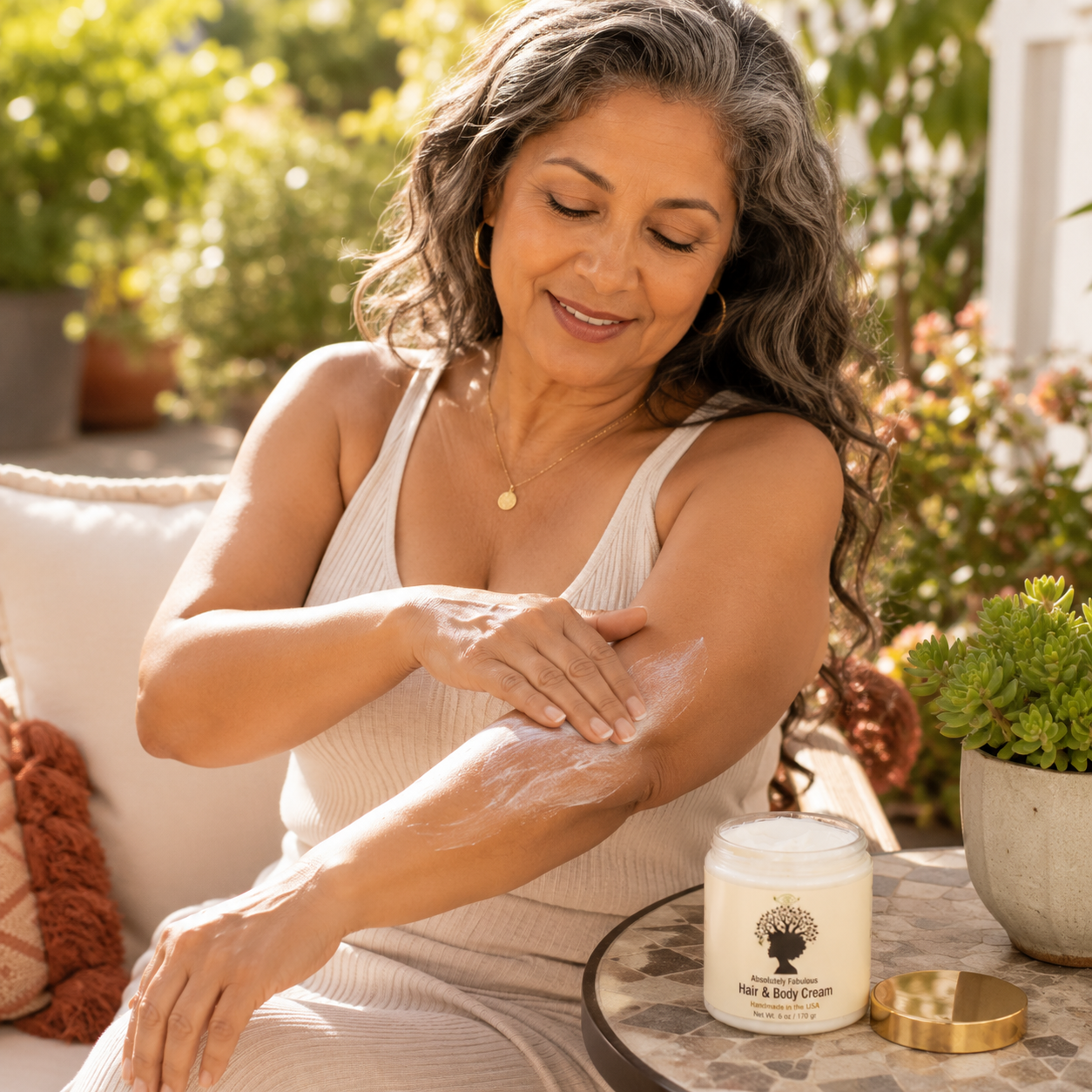 Latina woman in her 50s with silver-streaked wavy hair applying Absolutely Fabulous Hair & Body Cream to her forearm while seated on a sunlit patio with the cream jar displayed on a small outdoor table.