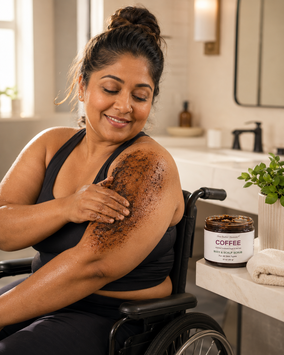 South Asian woman in her 40s using a wheelchair applying Shea Butter Heaven Coffee Body & Scalp Scrub to her shoulder in a bright accessible bathroom with warm natural light and the scrub jar displayed nearby.