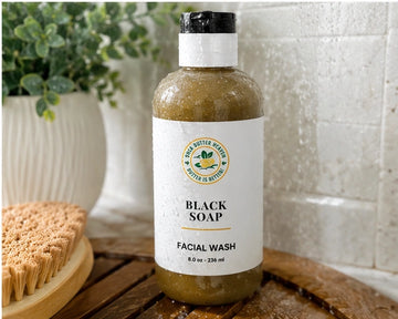 Shea Butter Heaven Black Soap Facial Wash in a shower setting with water droplets, greenery, and a natural body brush.